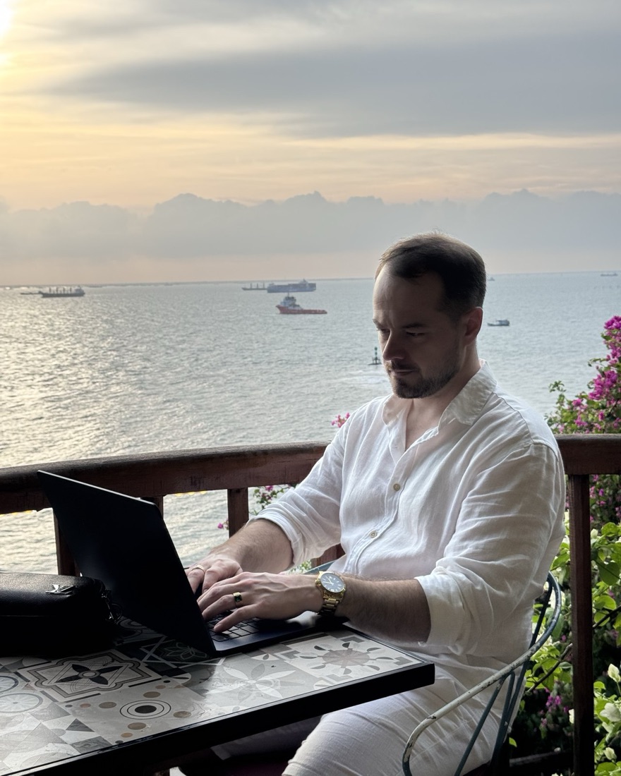 Blake Blackwell typing on a laptop at a balcony table above a harbor, ships on the water.