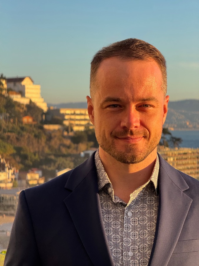 Blake Blackwell, smiling, in a navy blazer on a balcony above the sea at golden hour.